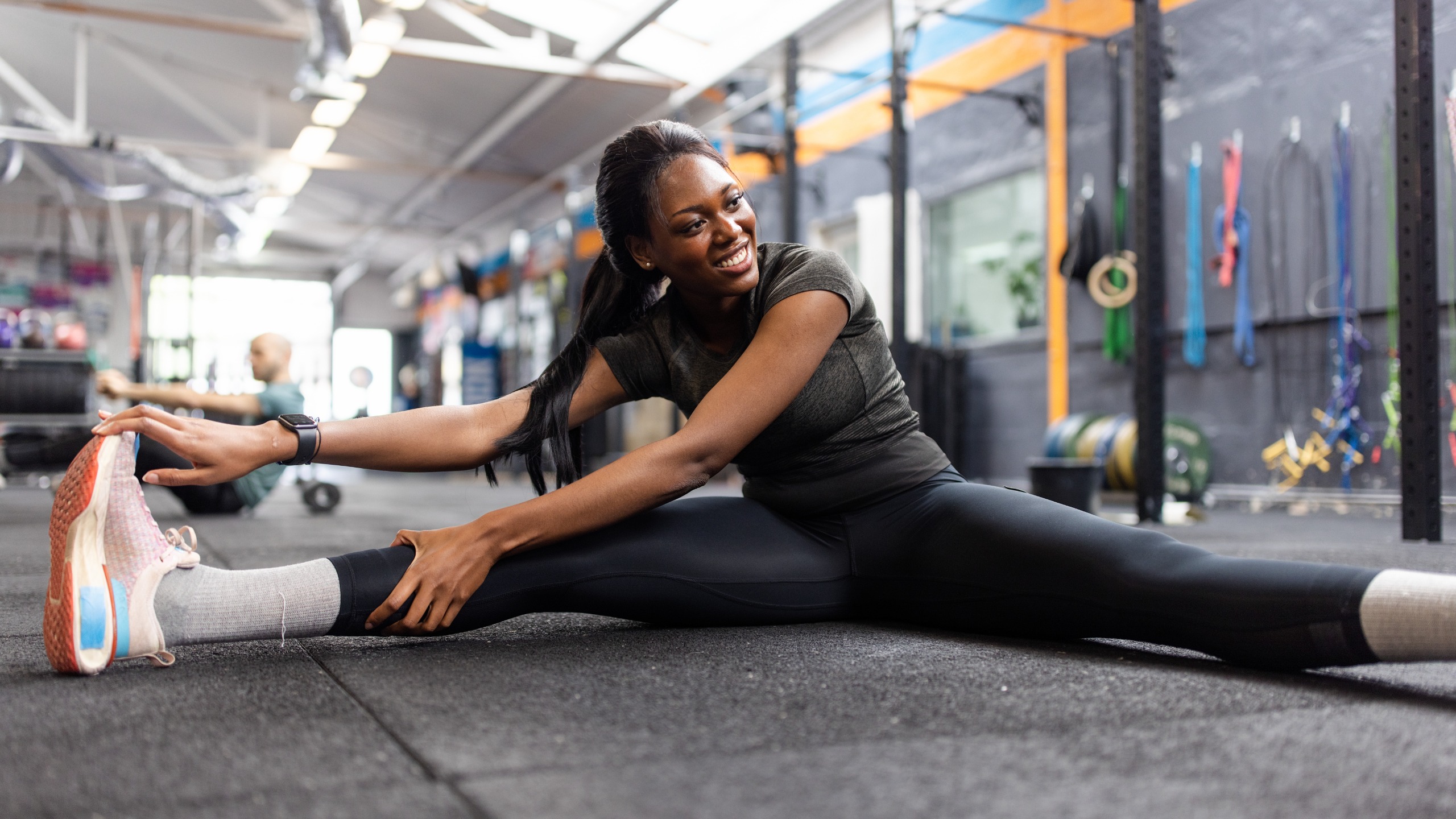A woman sits on the floor of her gym with her straight legs out wide. She leans towards her right leg and holds the top of her right sneaker in her hand.