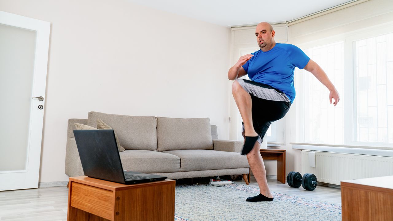 Man exercises in living room with a laptop open in front of him. He wears black and silver sports shorts and a blue T-shirt. One knee is raised to hip height.