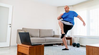 Man exercises in living room with a laptop open in front of him. He wears black and silver sports shorts and a blue T-shirt. One knee is raised to hip height.