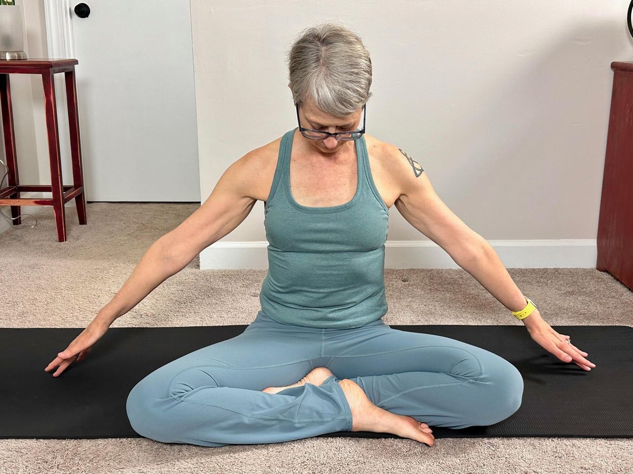 woman sitting cross-legged reaching her arms to the side on a black yoga mat in a living room