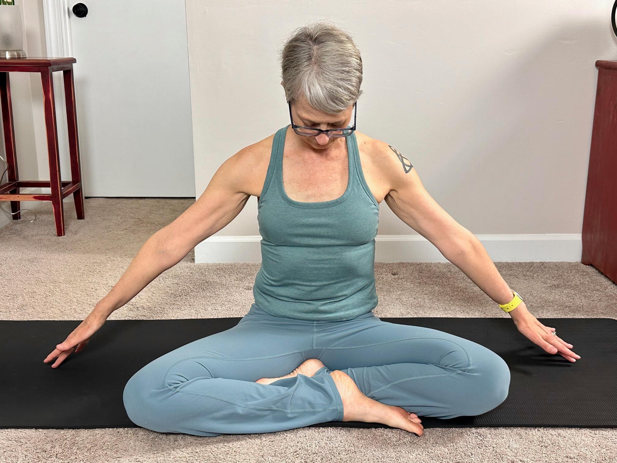 woman sitting cross-legged reaching her arms to the side on a black yoga mat in a living room