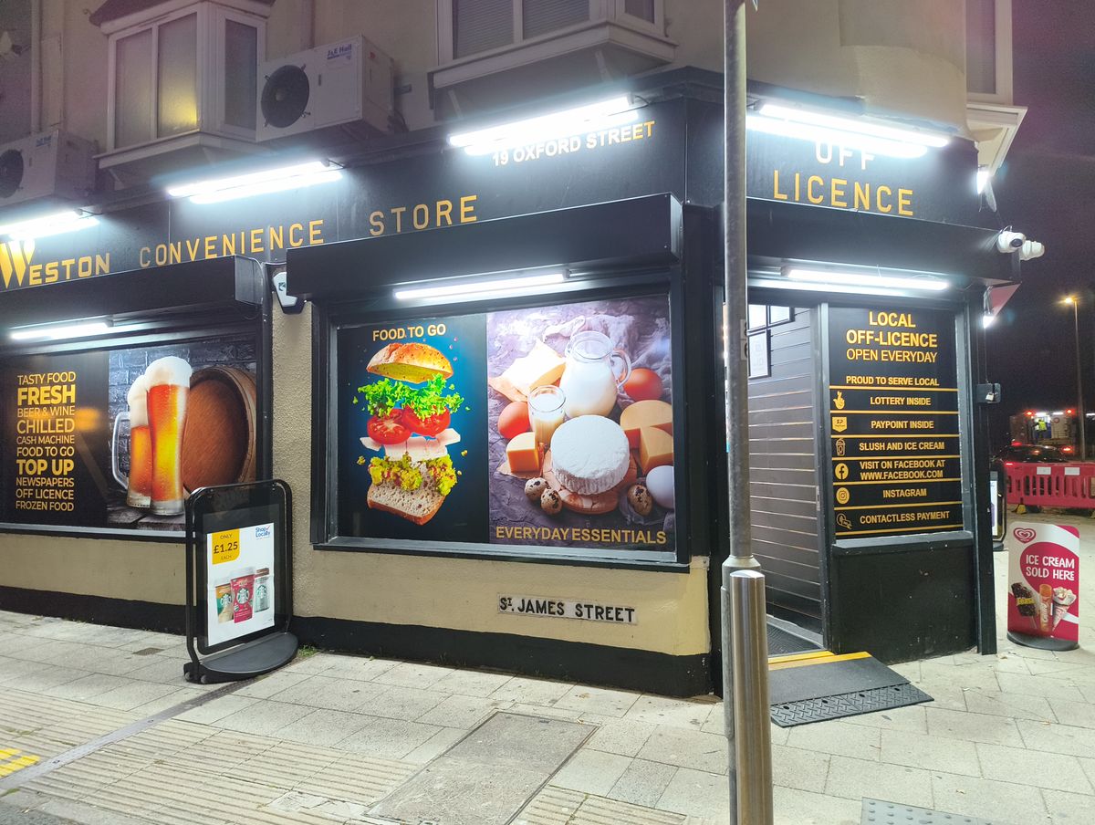 The exterior of the "Weston Convenience Store &amp; Off Licence" at night, featuring large window advertisements for food and everyday essentials.