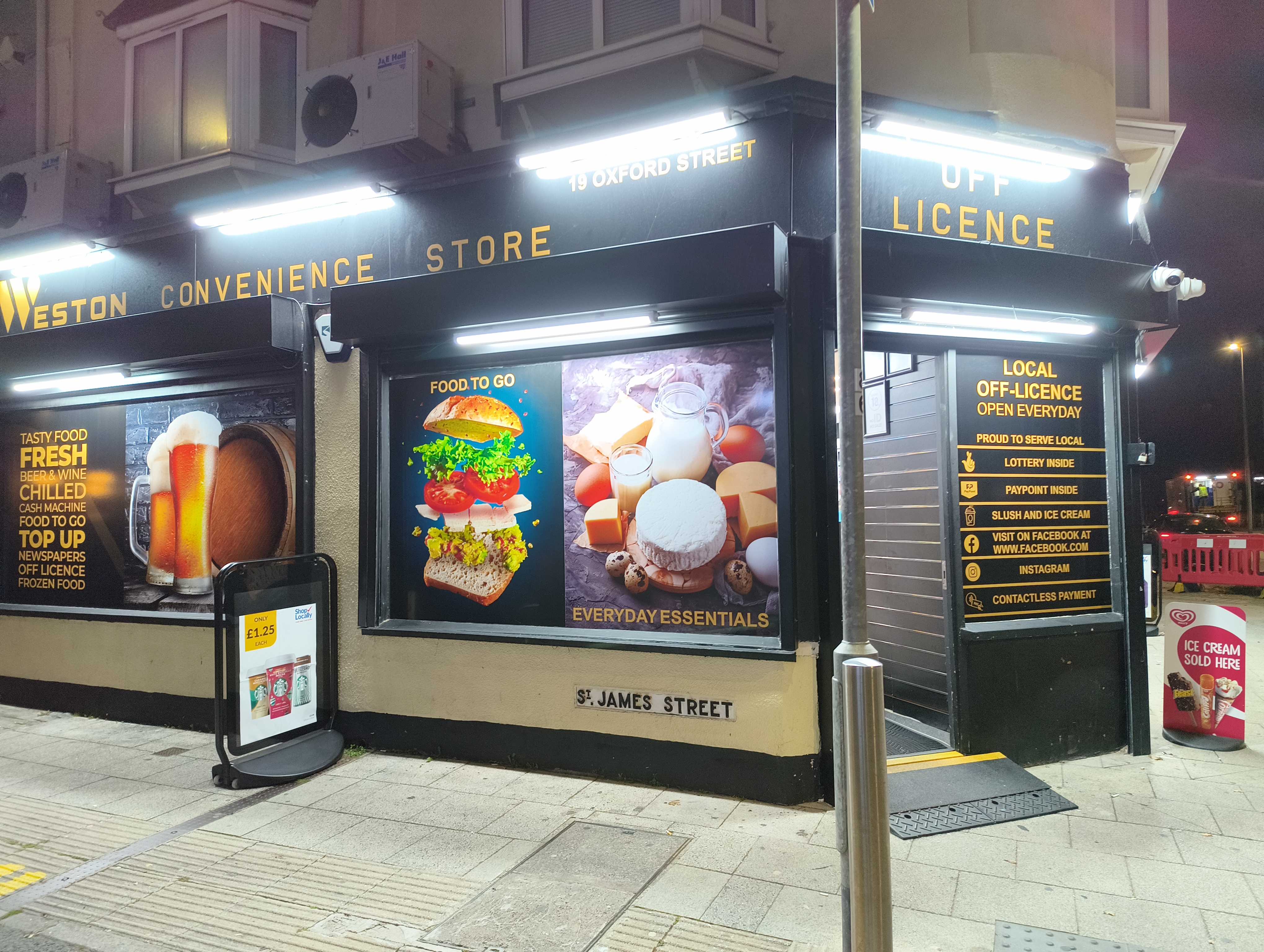 The exterior of the "Weston Convenience Store & Off Licence" at night, featuring large window advertisements for food and everyday essentials.