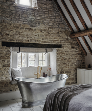 modernised bathroom in cotswold cottage with stone wall and aluminium freestanding bath and visiblew wooden beams on ceiling