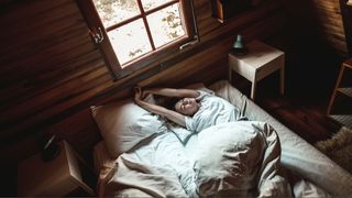 A woman laying on her back in bed with white sheets stretching her arms overhead in wooden cabin