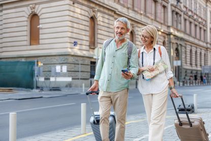 A senior couple uses phone while carrying luggage in a city.