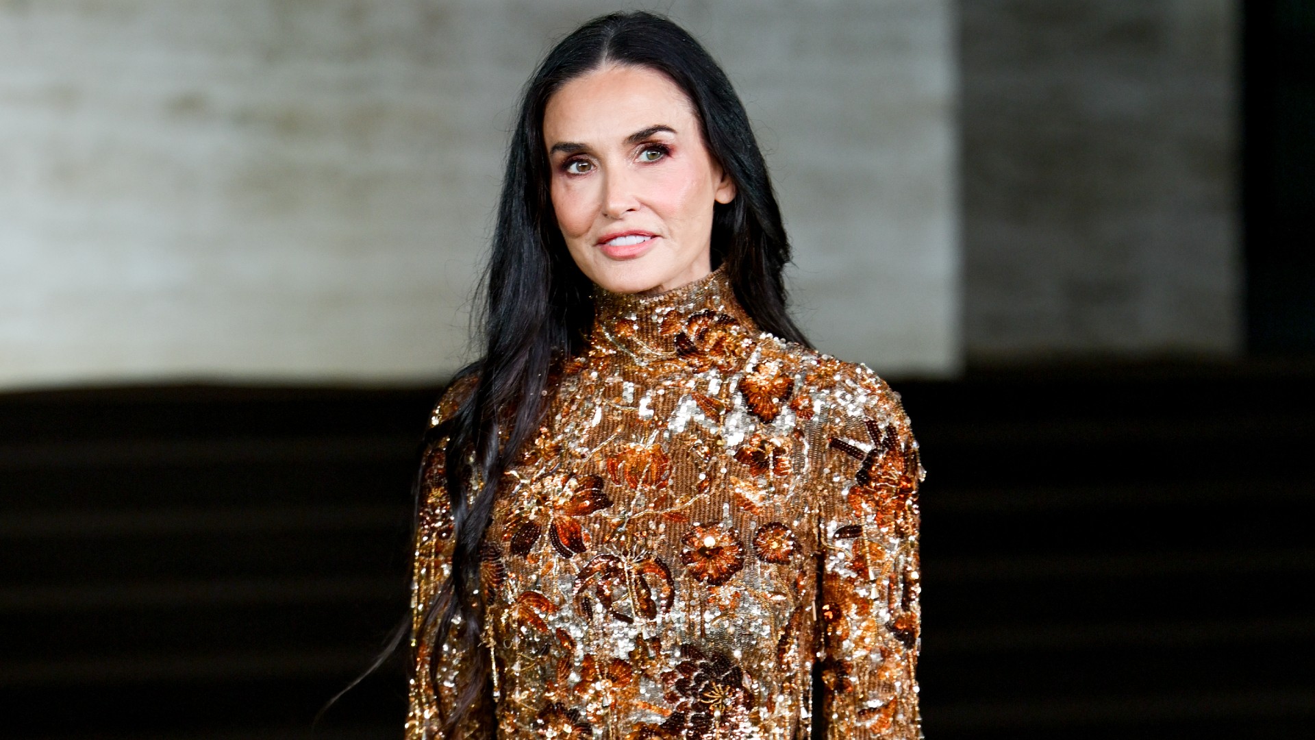 a white woman with long dark hair wearing a long-sleeve gold gown with beads and sequins