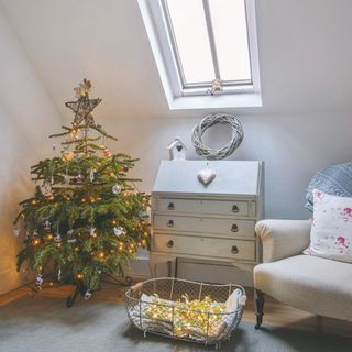 Small Christmas tree decorated with fairy lights, ornaments and a star topper next to drawer cabinet, chair and basket of fairy lights