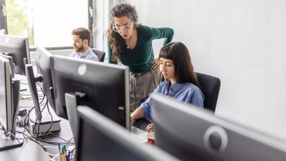 Male and female software developers working on desktop computers in an open plan office space with senior employee leaning over and observing work.