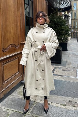 spring trench coats shown in a photo of a French woman standing outside on the sidewalk in Paris wearing aviator sunglasses, a tan funnel neck trench coat, black leggings, black pointed pumps, and a black clutch bag