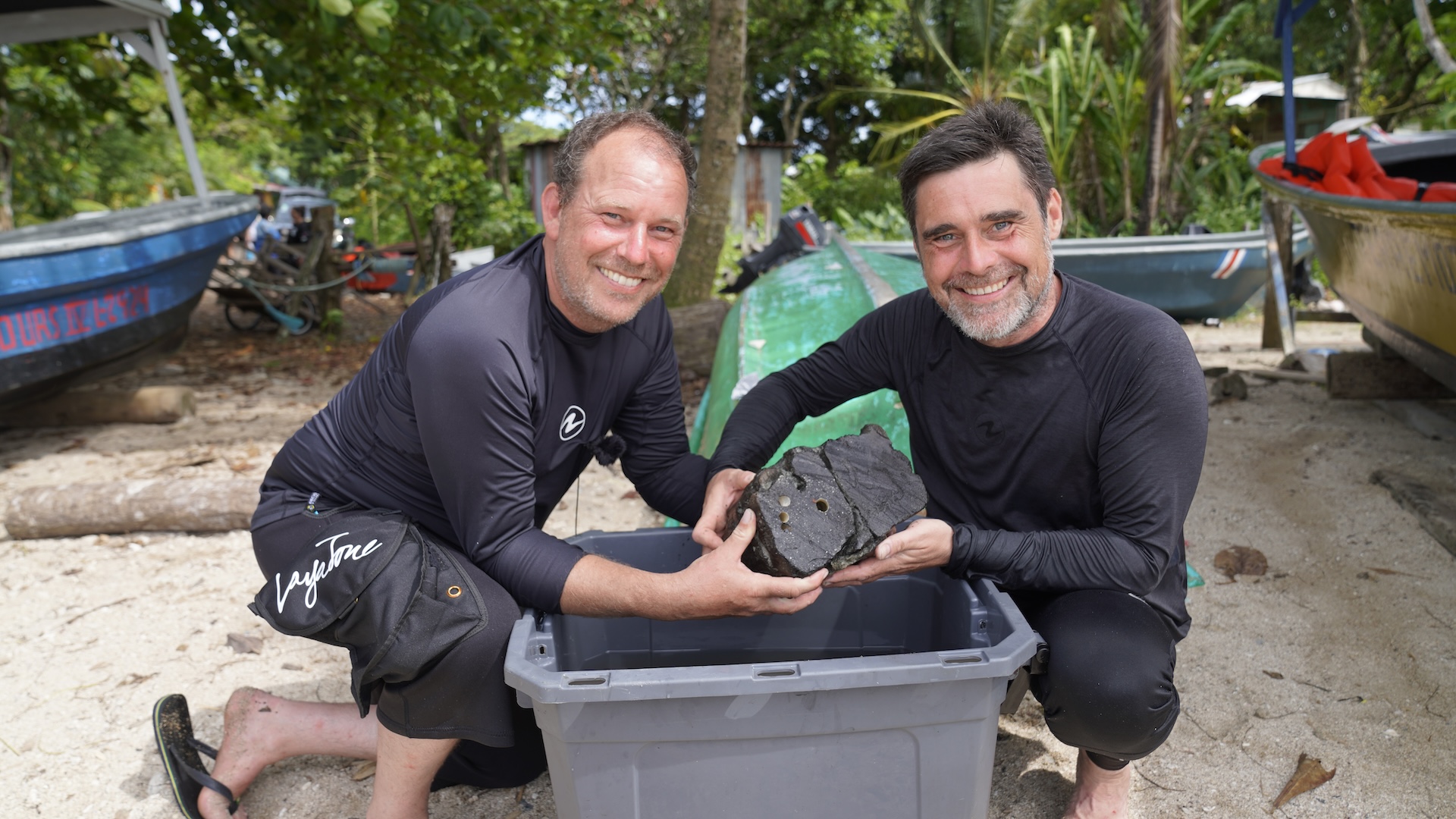 Two men hold up excavated ship timbers on a beach