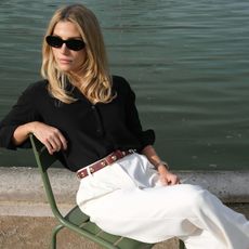 Blonde woman wearing black button down shirt, white linen pants, brown belt, and black sunglasses sits on park bench in front of a lake.