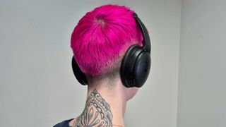 A woman with short pink hair is wearing a pair of black Bose QuietComfort headphones.