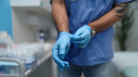 A man wearing a brown watch, blue shirt and jeans puts on a blue pair of nitrile gloves in a blurry hospital room in the background