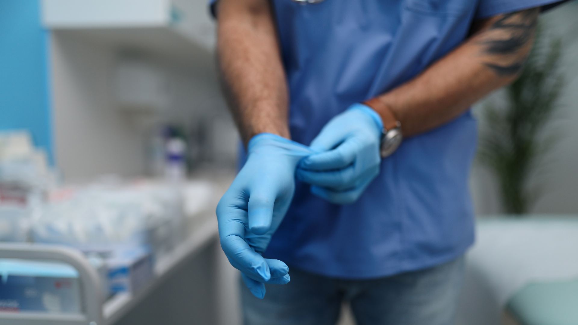A man wearing a brown watch, blue shirt and jeans puts on a blue pair of nitrile gloves in a blurry hospital room in the background