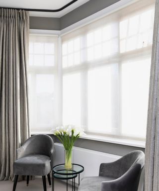 Two dark grey armchairs in a bay window with net curtains and grey curtains.