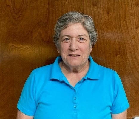 Headshot of woman with short gray hair in a blue polo shirt on dark wooden background