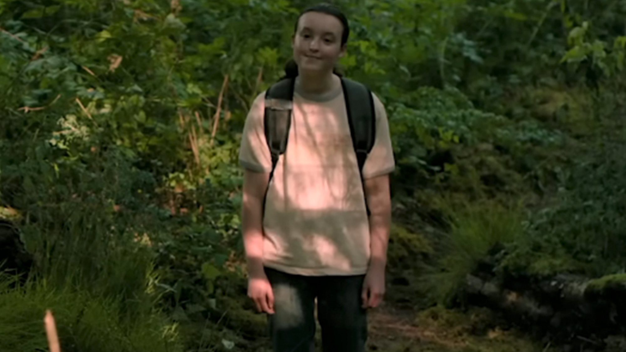 Bella Ramsey wearing a white tee-shirt and backpack in the woods in The Last Of Us season 2