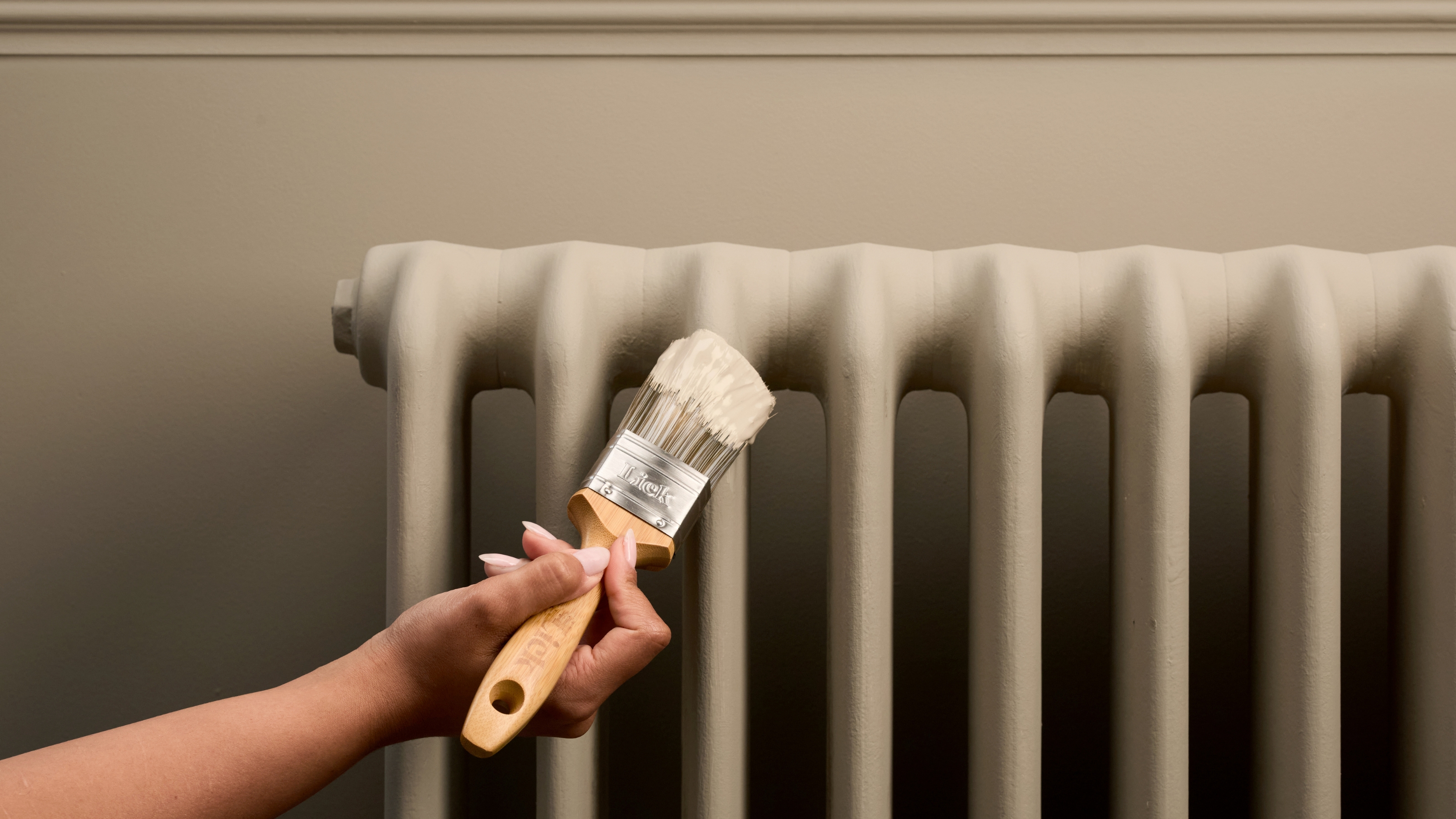 Image of an arm holding a paint brush that is painting a radiator and wall in the same matt, beige paint color. 