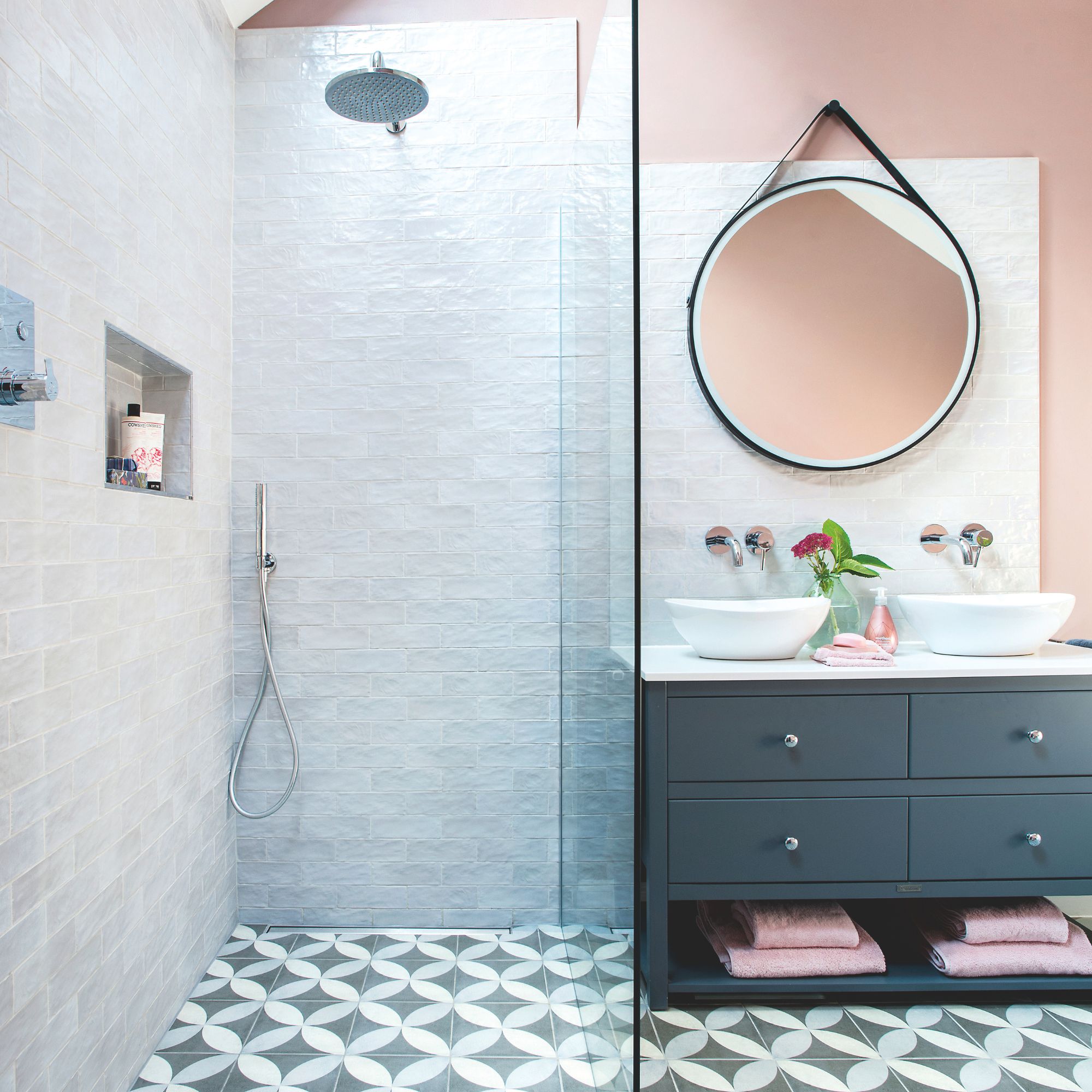 Walk-in shower with glass screen next to a double vanity with two sinks and circular mirror above