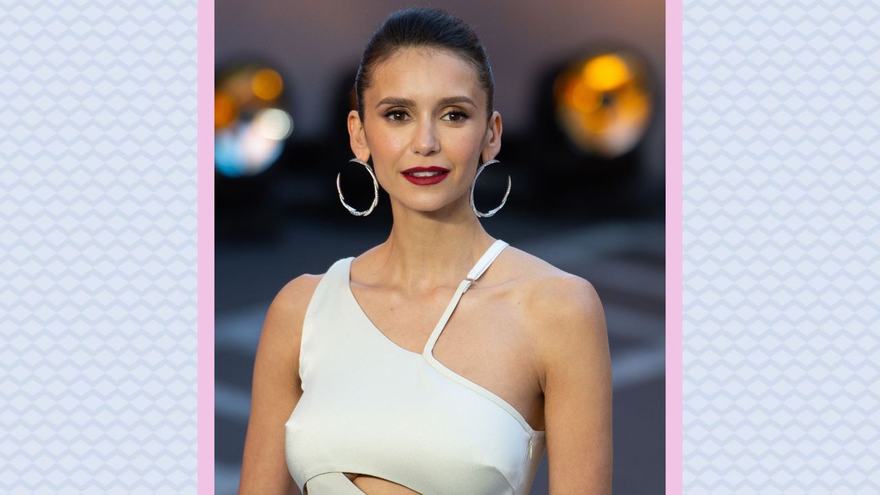 Nina Dobrev smiling, wearing a one shoulder white dress as she attends the Royal Performance of &quot;Top Gun: Maverick&quot; at Leicester Square on May 19, 2022 in London, England. / on a blue and pink background