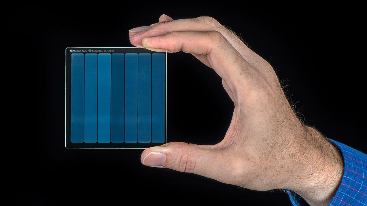 A hand holding a .5 cm x 7.5 cm x 2 mm piece of silica glass used for glass data storage against a black background.