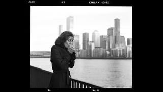 A figure in a black coat stands by a railing, overlooking a city skyline reflected in the water on a cloudy day