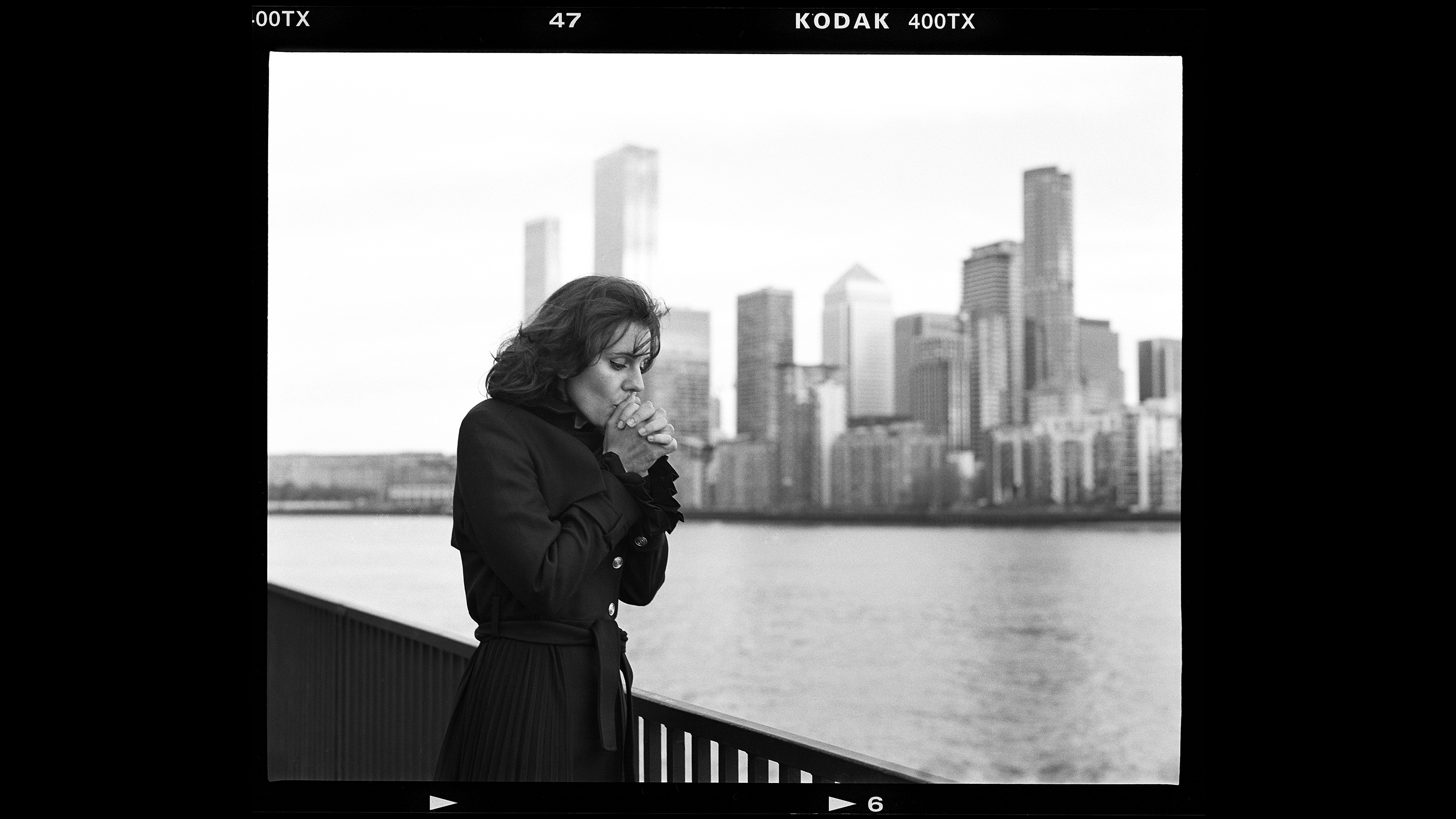 A figure in a black coat stands by a railing, overlooking a city skyline reflected in the water on a cloudy day