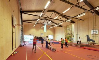 Gymnasium in the KIEM building, high ceiling with black metal beams, strobe lighting, neutral walls, red floor marked out with sport lines, adult supervisors, children playing sport games, sports equipment, windows and doorways