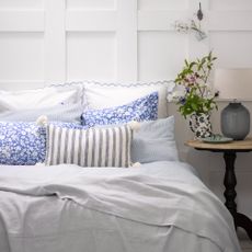 A panelled bedroom with a bed covered in pillows and cushions and a sheet instead of duvet with a bedside table next to it