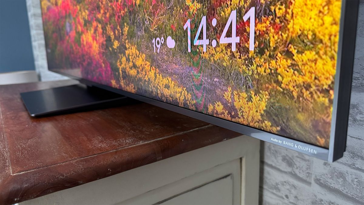 A close-up of the bottom corner of the 65-inch TCL C7K Mini LED TV on a TV cabinet in front of a brick wall. Flowers are partially visible on screen.