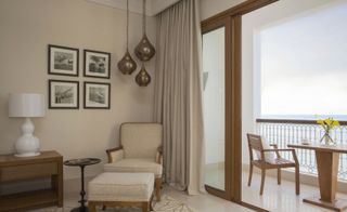 Hotel room balcony with wooden chair and table, cream armchair with ottoman and cream curtains