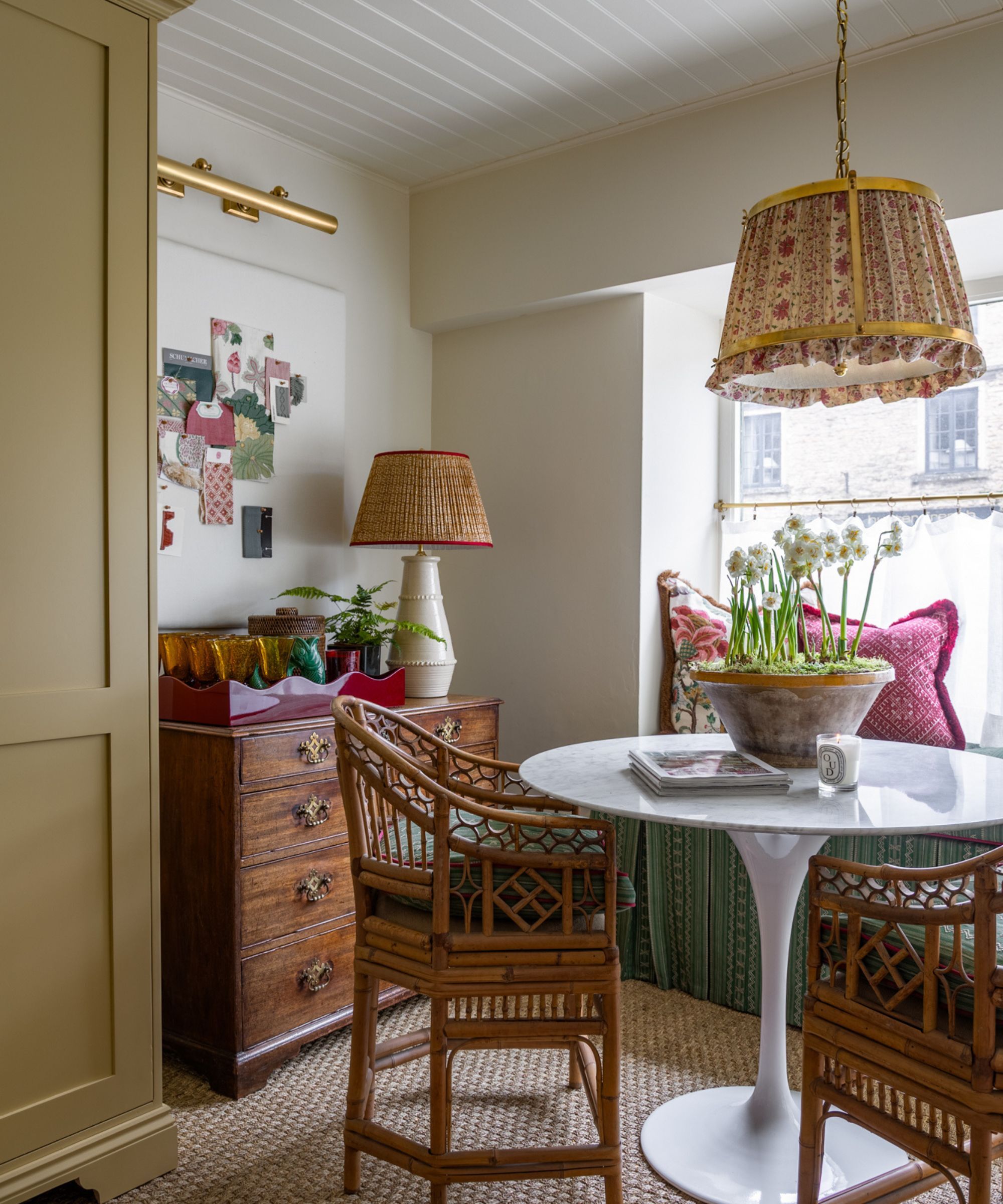 A corner of a room with a chest of drawers, on top is a pretty lamp and decorative items. There is a white gloss circular table with two bamboo style chairs. On top of the table is a Diptyque candle and a rustic bowl with daffodils planted in moss. Sisal carpet on the floor. Neutral walls.