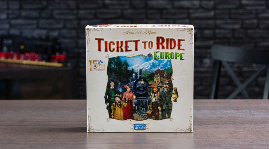 Förpackningen till sällskapsspelet Ticket To Ride står på ett bord framför en grå tegelvägg.