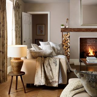 autumnal living room with neutral scheme, patterned curtains and cushions, log pile, stool with lamp, lit open fire