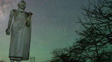 A large statue is seen with a meteor shower behind it in the night sky