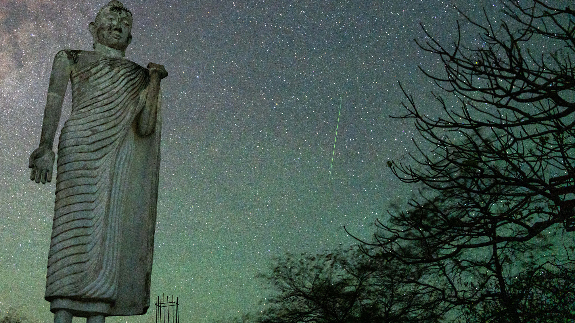 A large statue is seen with a meteor shower behind it in the night sky