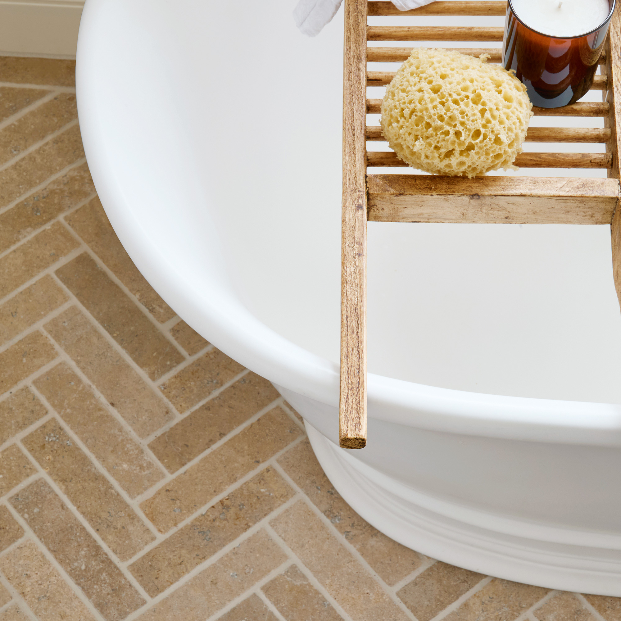a neutral bathroom with a large white freestanding bath tub and limestone herringbone floor tiles