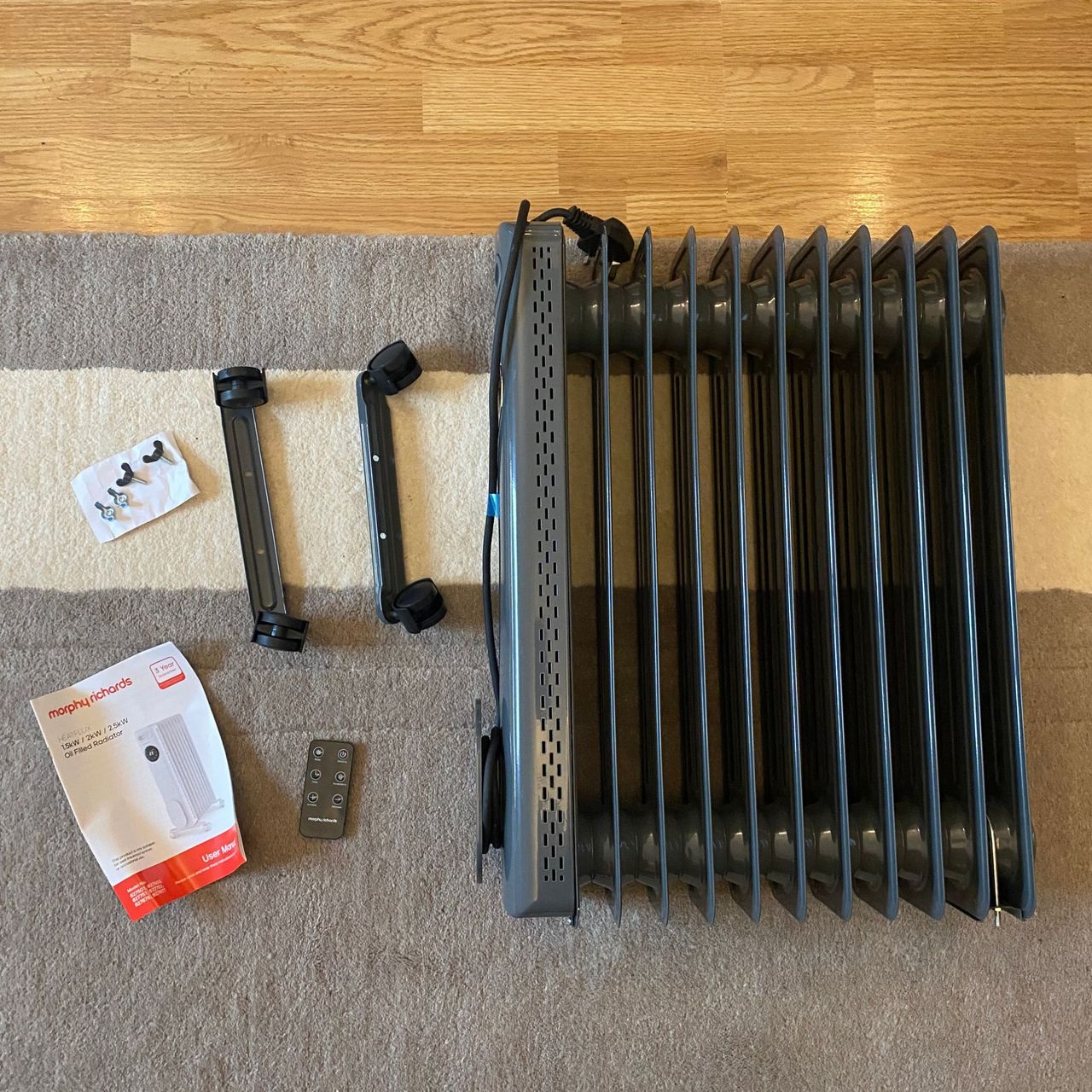 Flat lay of the Morphy Richards HeatFlux 2.5kW Digital Oil-filled Radiator, wheel bases, remote control, bolts and instruction manual