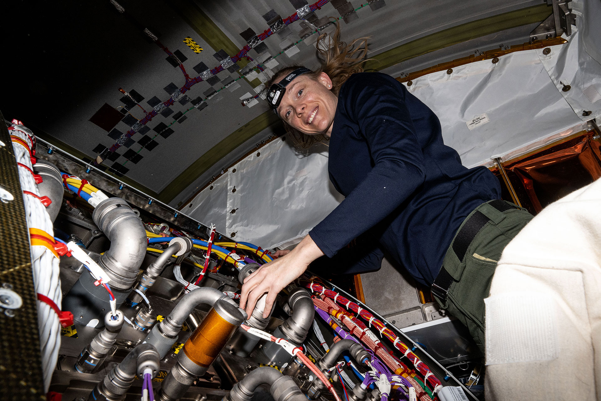 a woman wearing a dark blue long sleeve shirt, forest green pants and a headband with a light works near a panel of pipes and wires aboard a space station