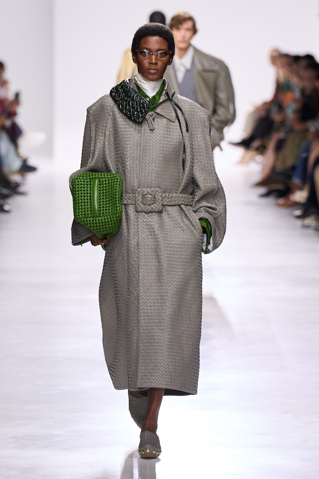 Layering staples for spring 2026 is shown in a photo from Bottega Veneta's runway show, with a model walking while wearing a woven gray leather trench coat, a black leather scarf around the neck, and a satin white turtleneck peeking out beneath the scarf and coat. The rest of the look includes silver wireframe glasses, a green clutch bag, and gray clogs