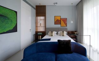 A bedroom with double bed and a blue sofa at the end of the bed. above the head of the bed are two abstract frames haning on a grey wall.