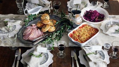 A Thanksgiving, Christmas tablescape for a dinner party, with a white table runner on a brown wooden table, sliced ham, pastries, red cabbage, vintage plates, white napkins, silver flatware, and wine glasses filled with red wine.