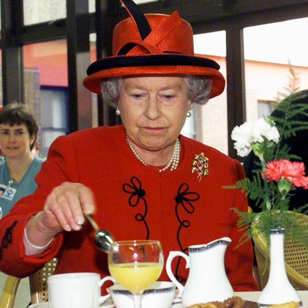 Queen Elizabeth wearing a red coat and hat stirring tea