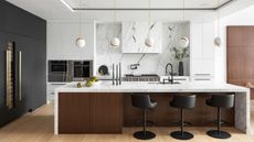 A kitchen with an island in the middle as well as bar stools beside it. There are five pendant lights hanging above the island. There is also a view of kitchen appliances as well well as a few kitchen accessories 