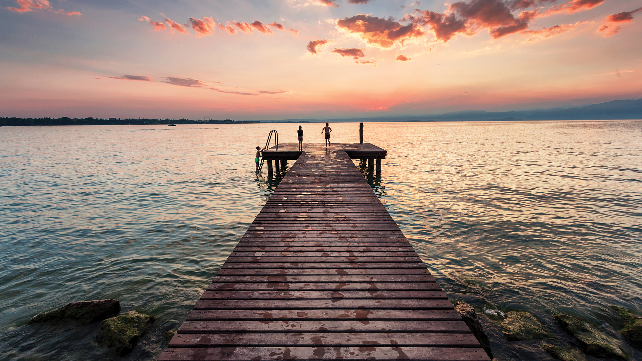 Two children play on a wooden pier at sunset, while another child splashes in the water, surrounded by tranquil waves and colorful skies