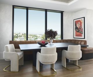 Modern dining space with large windows, dark table, fabric bench, and matching ivory upholstered chairs