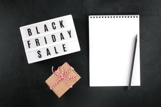 A black background with a notebook next to an LED sign that says Black Friday Sale, along with a brown paper package tied with red and white ribbon.