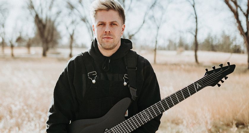 Mike Stringer wear a black hoodie and stands in a field of straw-colored long grass with trees in the background. He is holding an Aristides seven-string guitar.