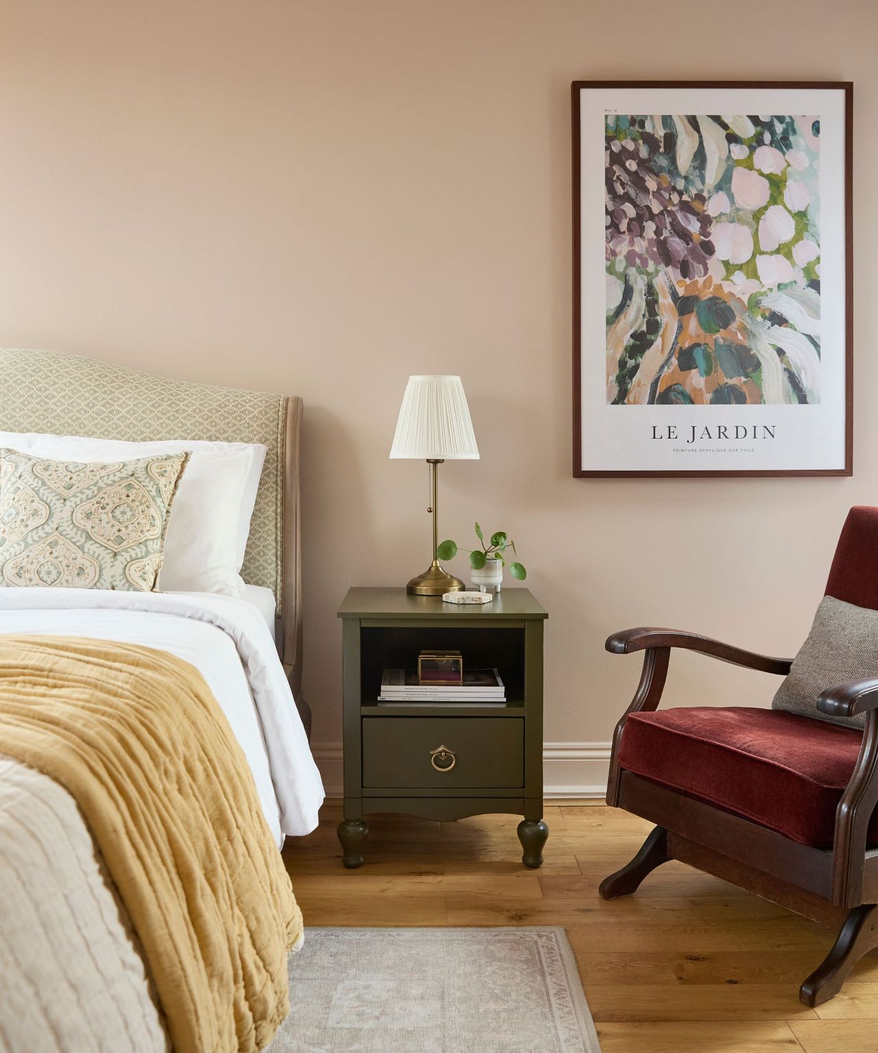 A bedroom with pale blush walls, a neutral toned bed, an olive green bedside cabinet, and a red chair